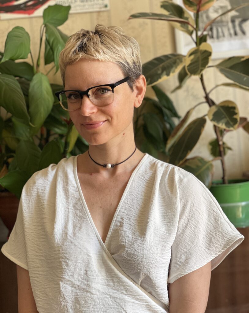 Portrait of Tatiana Vahan, wearing eyeglasses, a flow white blouse, and necklace sitting in front of plants in a sunlit room.